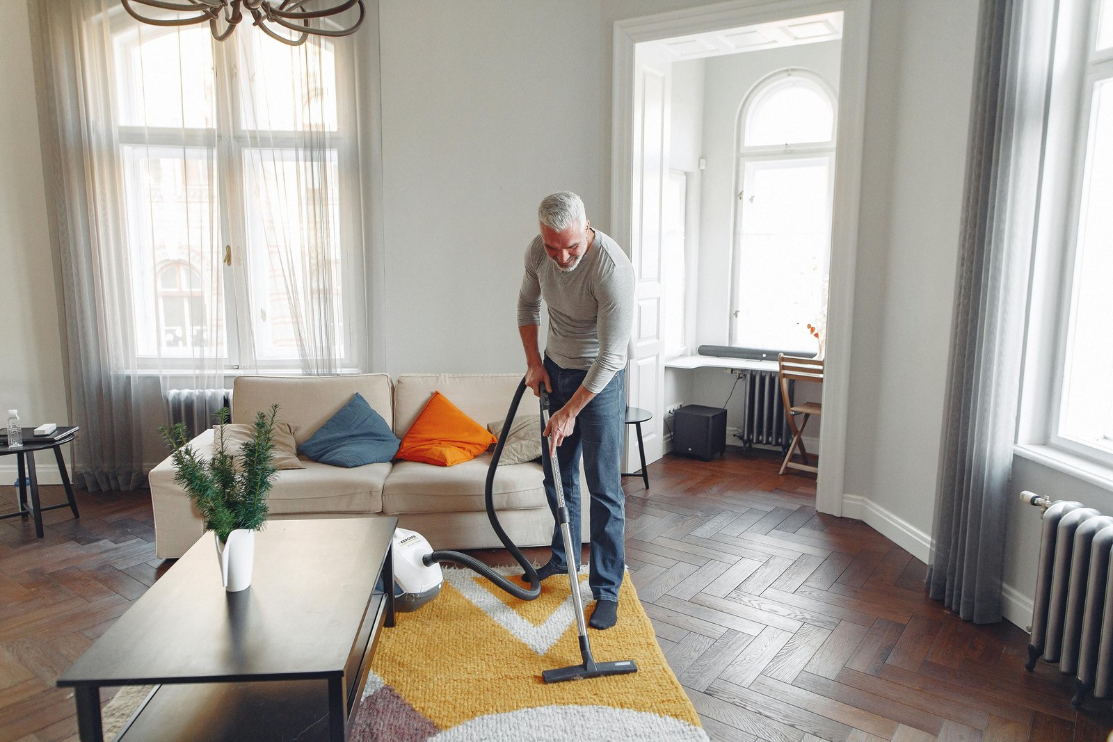 A senior man cleans the living room with a vacuum cleaner in a well-lit modern apartment.